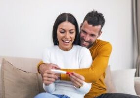 Enamoured couple finding out results of a pregnancy test sitting on the bed. Happy couple checking pregnancy test sitting on a couch in the living room at home. Family, parenting and medical concept